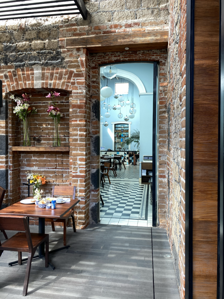 Interior view of Carlota restaurant in Mexico City, showing an exposed brick courtyard, arched brick niche, wooden dining table, and doorway leading into a tiled dining room. The space blends historic brick architecture with graphic black-and-white flooring, modern lighting, and a relaxed Mexico City restaurant atmosphere.