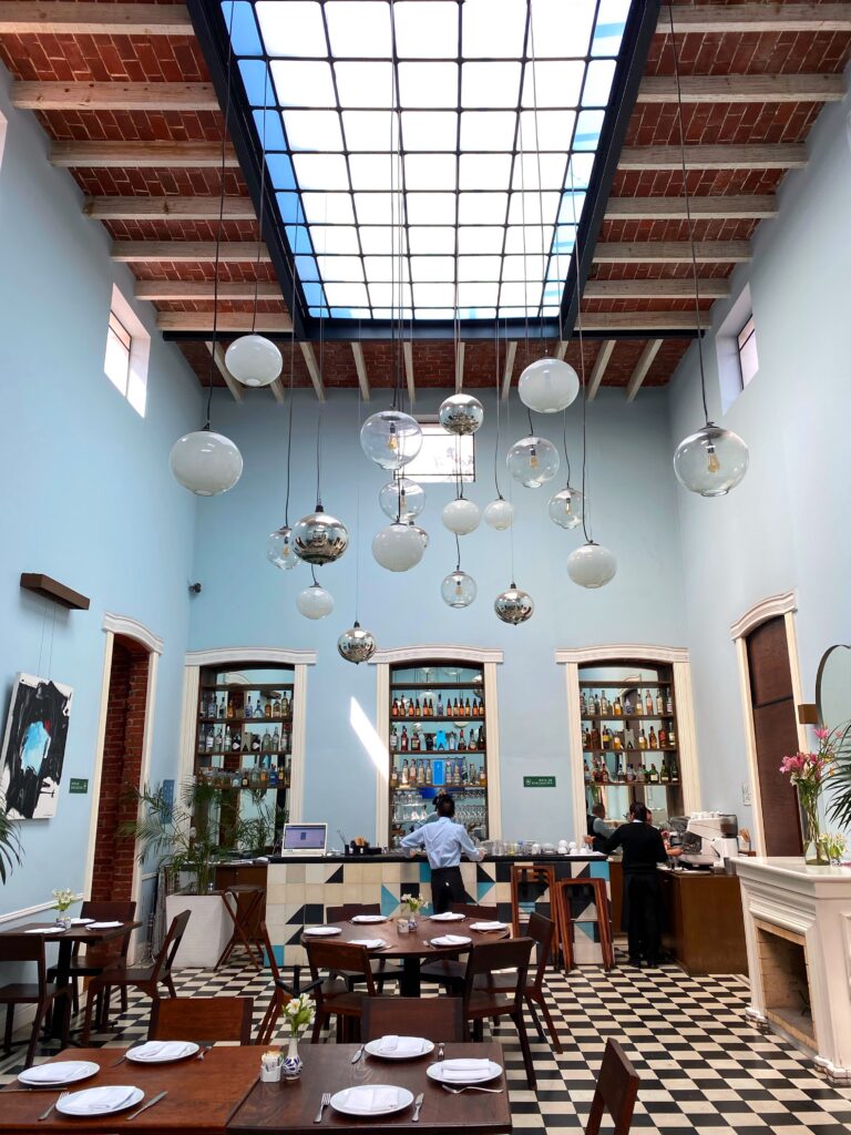 Interior of Carlota restaurant in Mexico City, now closed, with a dramatic skylight, exposed brick ceiling, hanging globe lights, pale blue walls, and black-and-white checkered tile floors. The airy dining room features wood tables, a tiled bar, and historic architectural details that give the former Mexico City restaurant a polished, design-rich atmosphere.