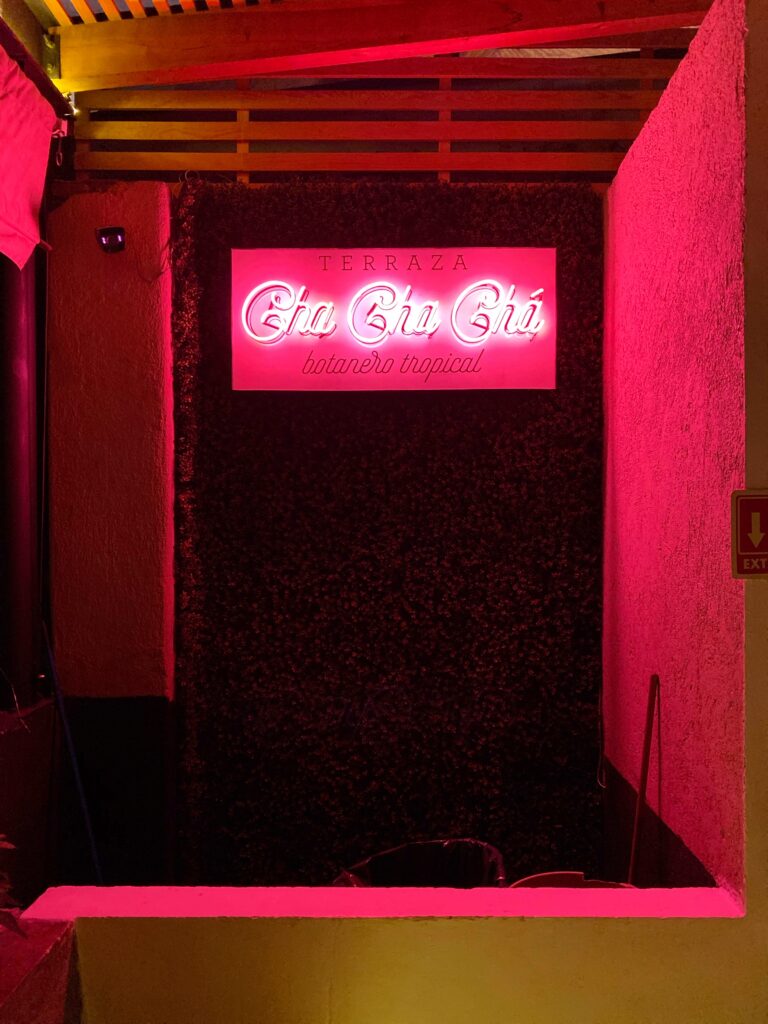 Neon sign for Cha Cha Chá restaurant in Mexico City, glowing pink against a dark greenery wall on the terrace entrance. The bold lighting and tropical botana-style branding create a playful, high-energy Mexico City dining moment.