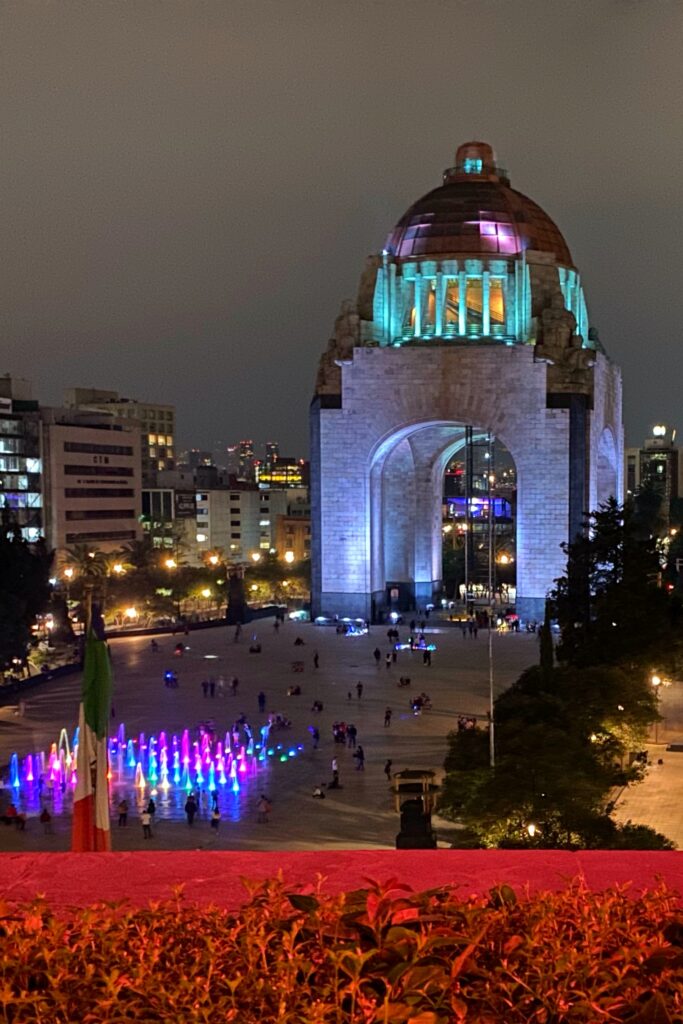 Night view from Cha Cha Chá restaurant in Mexico City overlooking Monumento a la Revolución, with the illuminated dome, city skyline, and colorful fountain lights in the plaza below. The terrace view adds a dramatic Mexico City backdrop to the dining experience, blending architecture, nightlife, and rooftop atmosphere.