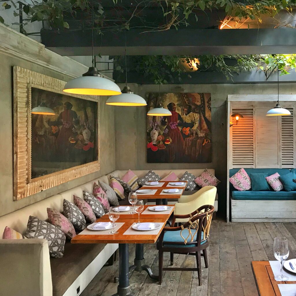 Interior of La Única in Mexico City with cream banquette seating, patterned pillows, wooden tables, pendant lights, and oversized framed artwork along the wall. The layered greenery, rustic wood floors, and colorful textiles create a warm, eclectic restaurant atmosphere with a relaxed Mexico City design sensibility.