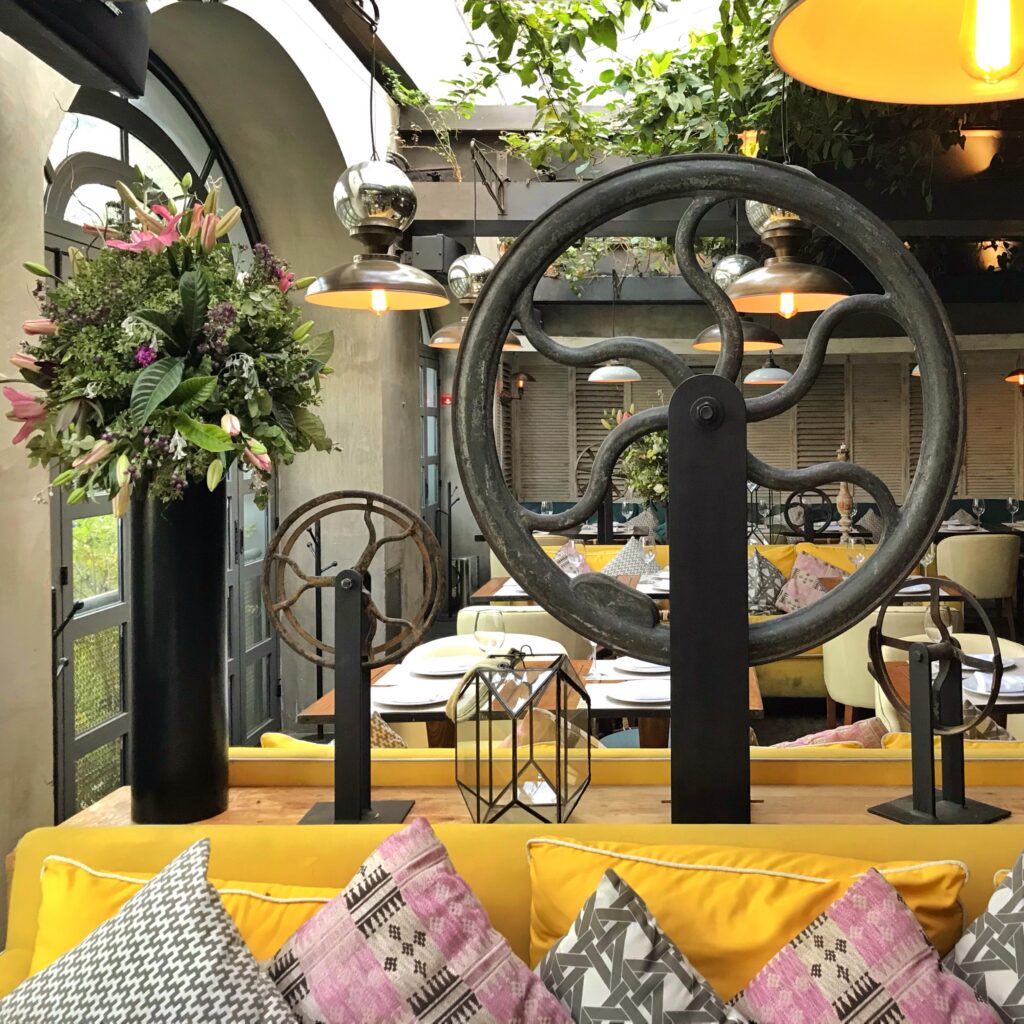 Interior of La Única in Mexico City with bright yellow banquette seating, patterned pillows, oversized industrial wheel sculptures, and lush greenery overhead. The restaurant mixes bold color, metal lighting, arched windows, and eclectic decor for a lively, design-forward dining atmosphere.