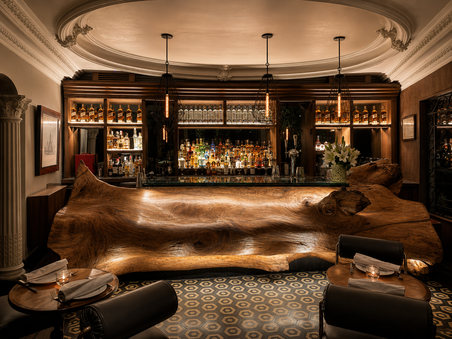 Interior of Sylvestre in Mexico City featuring a dramatic live-edge wood bar counter, backlit bottles, black cage pendant lights, and ornate ceiling molding. The moody restaurant bar blends carved wood, warm lighting, patterned tile, and historic architectural details for a refined Mexico City dining atmosphere.