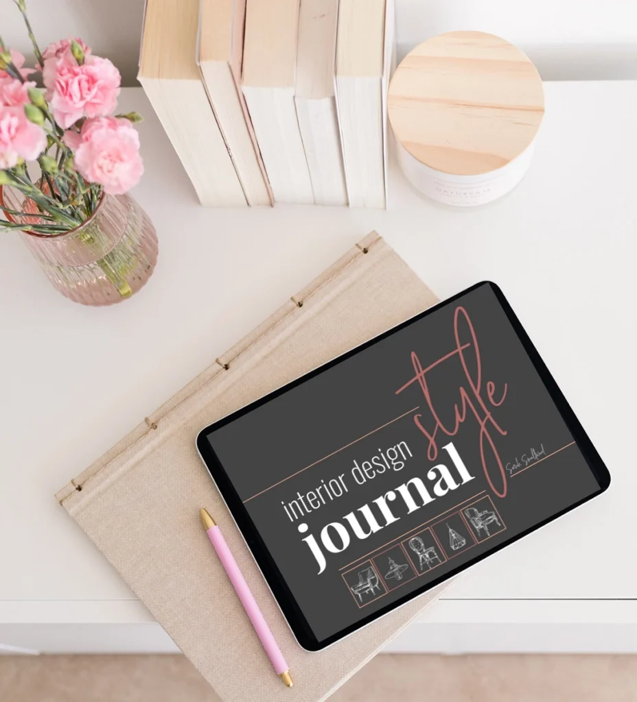 Flat lay of an interior design style journal displayed on a tablet beside a linen notebook, pink pen, books, candle, and fresh pink flowers. This image represents using a design journal to clarify your interior design style, organize inspiration, and create a more intentional plan for your home.