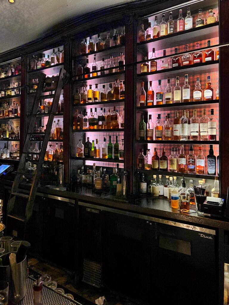 Back bar at 7 Grand in downtown Los Angeles with tall dark wood shelving, a rolling library ladder, and rows of illuminated liquor bottles. Pink backlighting, black cabinetry, and the densely stocked bottle display give the bar a moody, old-school cocktail lounge atmosphere.