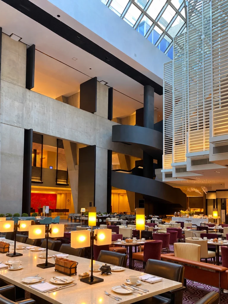 Lobby atrium at Hyatt Regency Mexico City with a large dining area, glowing table lamps, colorful chairs, and place settings arranged across long marble tables. The space features a dramatic black spiral staircase, concrete architectural walls, a glass ceiling, and warm lighting throughout the hotel lobby.