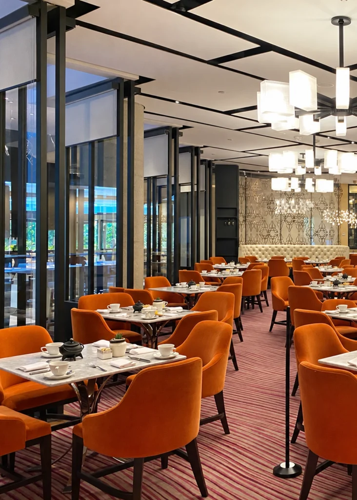 Mexico City restaurant dining room with bright orange upholstered chairs, marble-top tables, white place settings, and a pink striped carpet. Black-framed glass partitions, geometric ceiling details, cube pendant lights, and patterned wall panels give the space a polished, contemporary look.