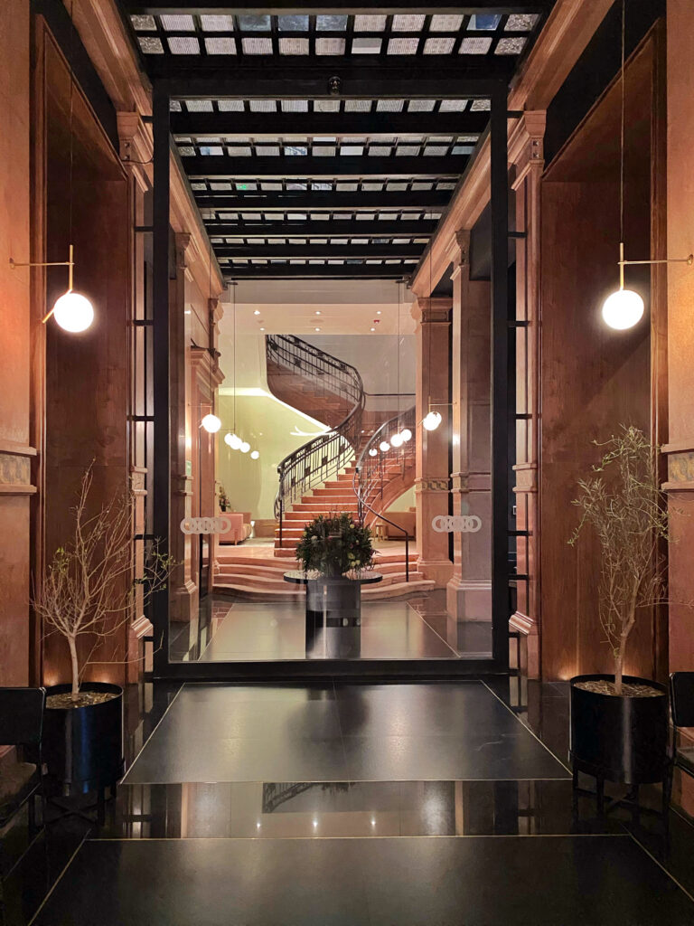Interior entry at Hotel Umbral Curio in Mexico City with glass doors, glossy black flooring, warm wood-paneled walls, and globe wall sconces. A curved staircase, black metal railings, potted branches, and a central floral arrangement are visible through the doorway.