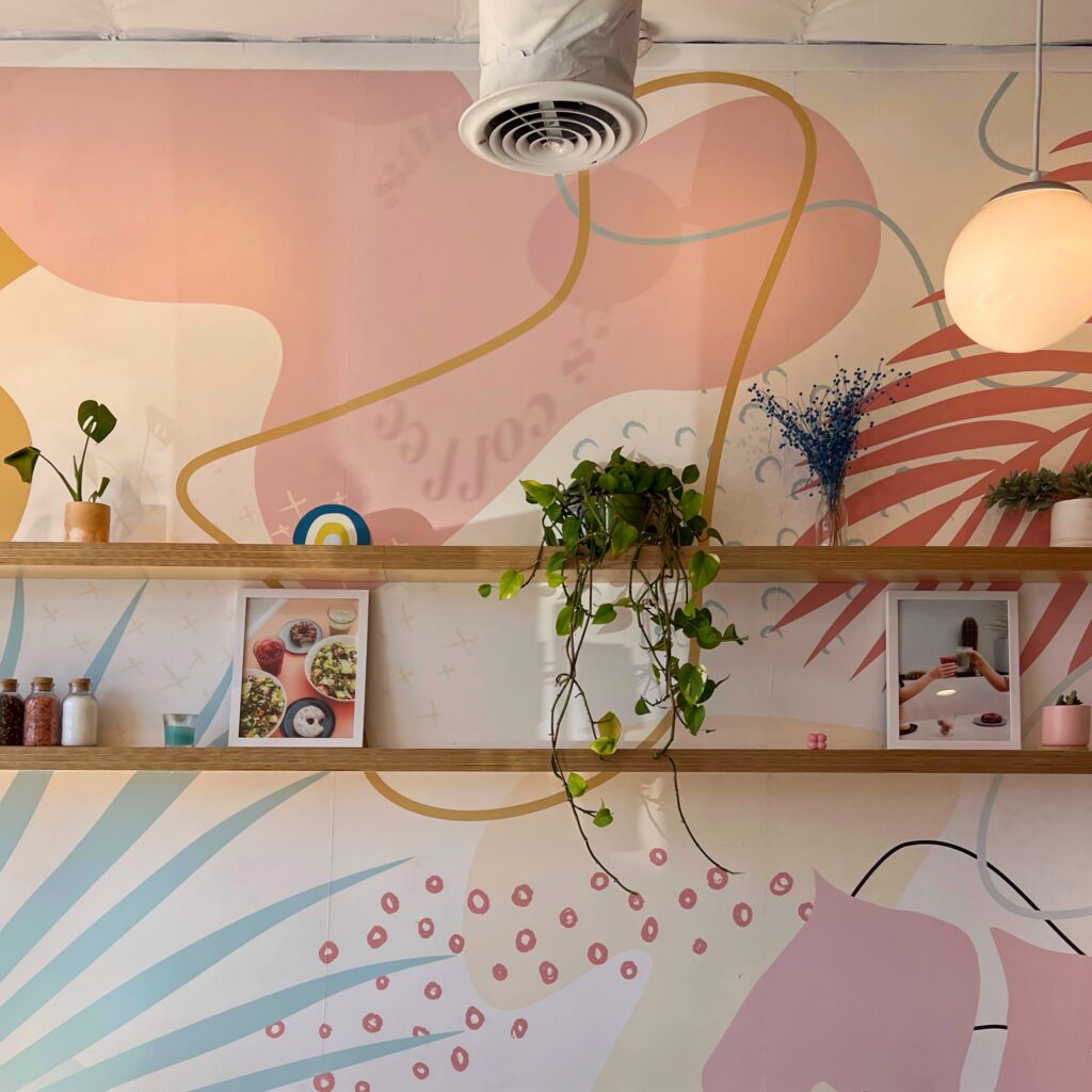 Interior of Alfalfa restaurant in Santa Monica with a pastel pink, blue, yellow, and coral abstract wall mural behind open wood shelves. Small plants, framed food photos, glass jars, and a glowing globe pendant add color and detail to the casual cafe space.