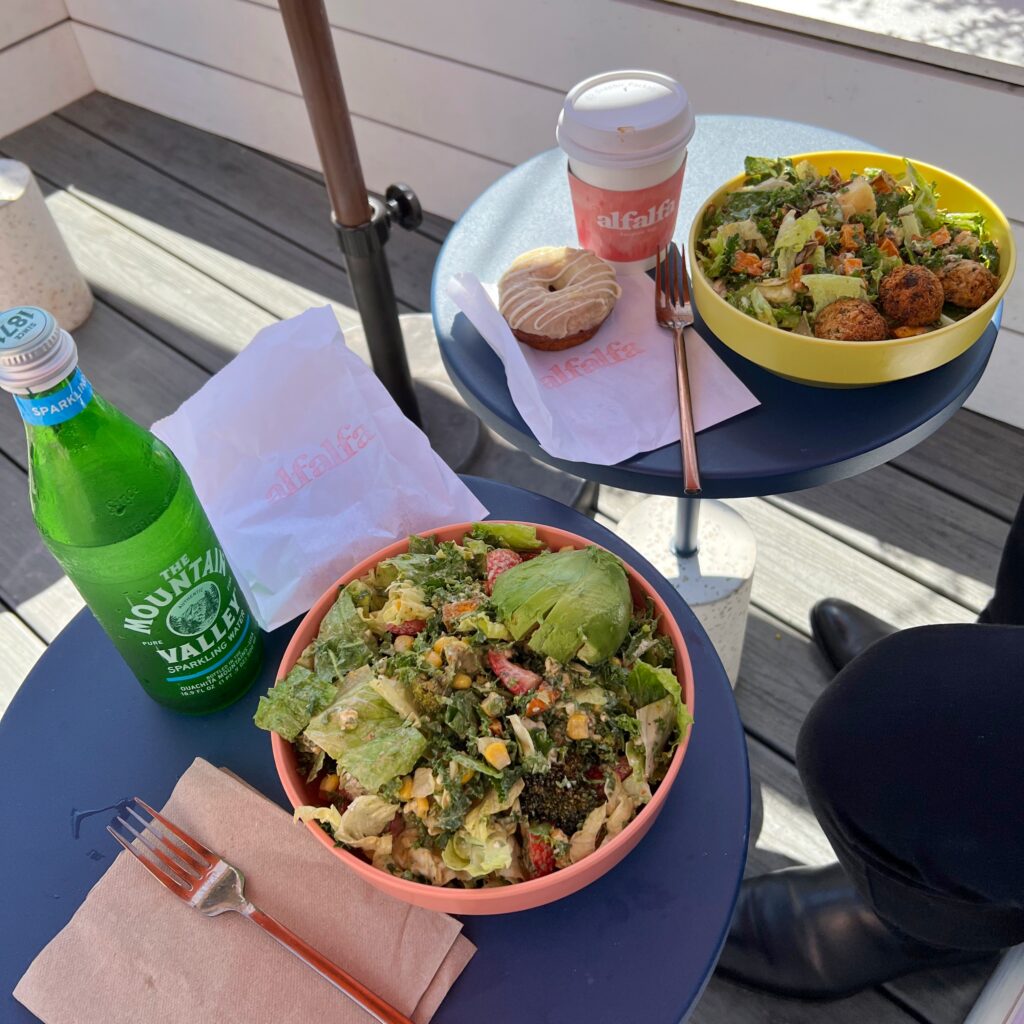 Outdoor patio table at Alfalfa in Santa Monica with two colorful salads, a coffee cup, a glazed donut, and a green bottle of Mountain Valley sparkling water. The food is served on small blue tables with Alfalfa-branded paper and bright bowls in the sunlight.
