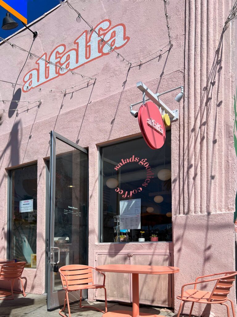 Exterior of Alfalfa restaurant in Santa Monica with a pale pink stucco facade, coral outdoor chairs, a matching patio table, and string lights casting shadows across the building. The storefront includes an open glass door, round Alfalfa sign, window graphics, and sunny sidewalk seating.