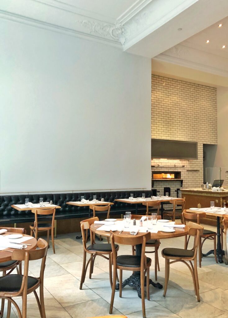 Dining room at Bottega Louie in Los Angeles with round wood tables, bentwood chairs, black tufted banquette seating, and a white brick fireplace. The space features tall white walls, ornate ceiling molding, marble floors, and a bright, airy restaurant interior.