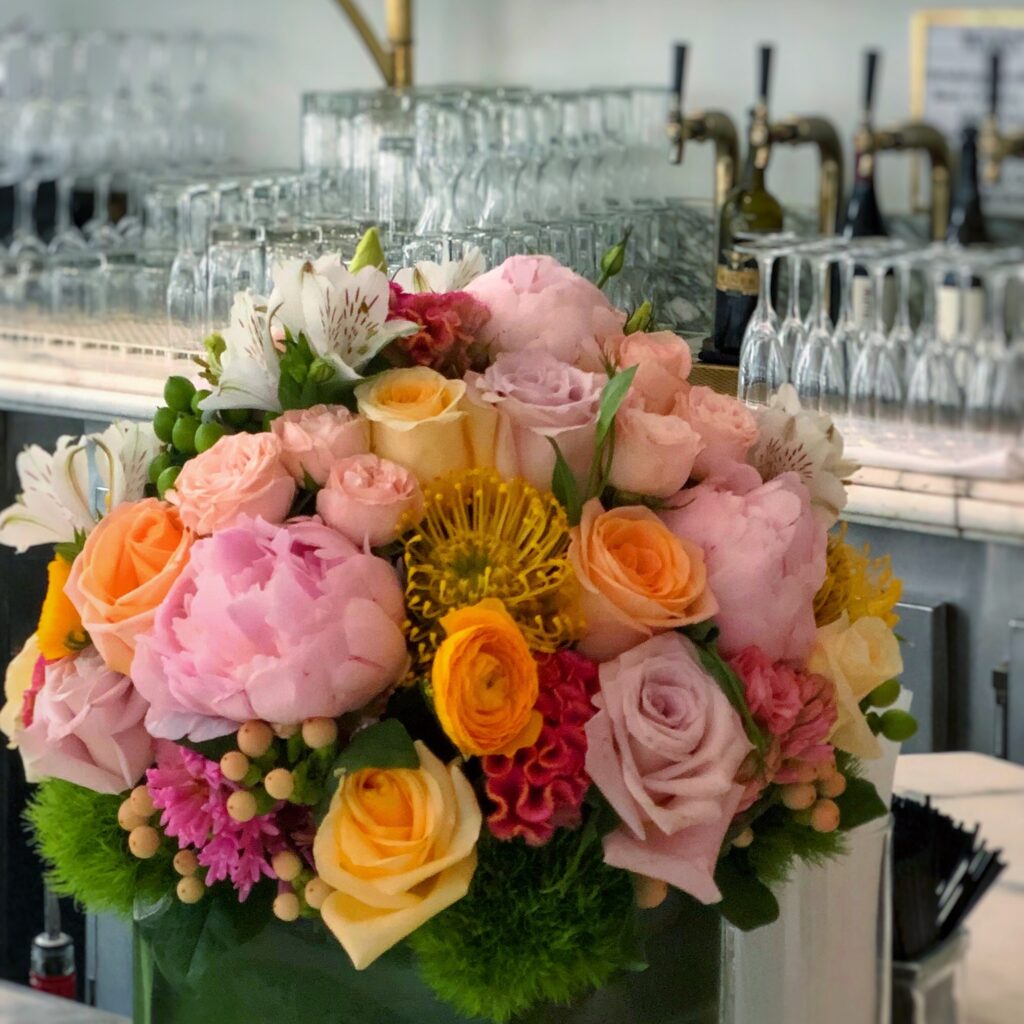 Colorful floral arrangement at Bottega Louie in Los Angeles with pink peonies, blush roses, orange roses, yellow flowers, white lilies, berries, and bright green accents. The bouquet sits in front of the restaurant bar with rows of glassware, wine bottles, and brass taps in the background.