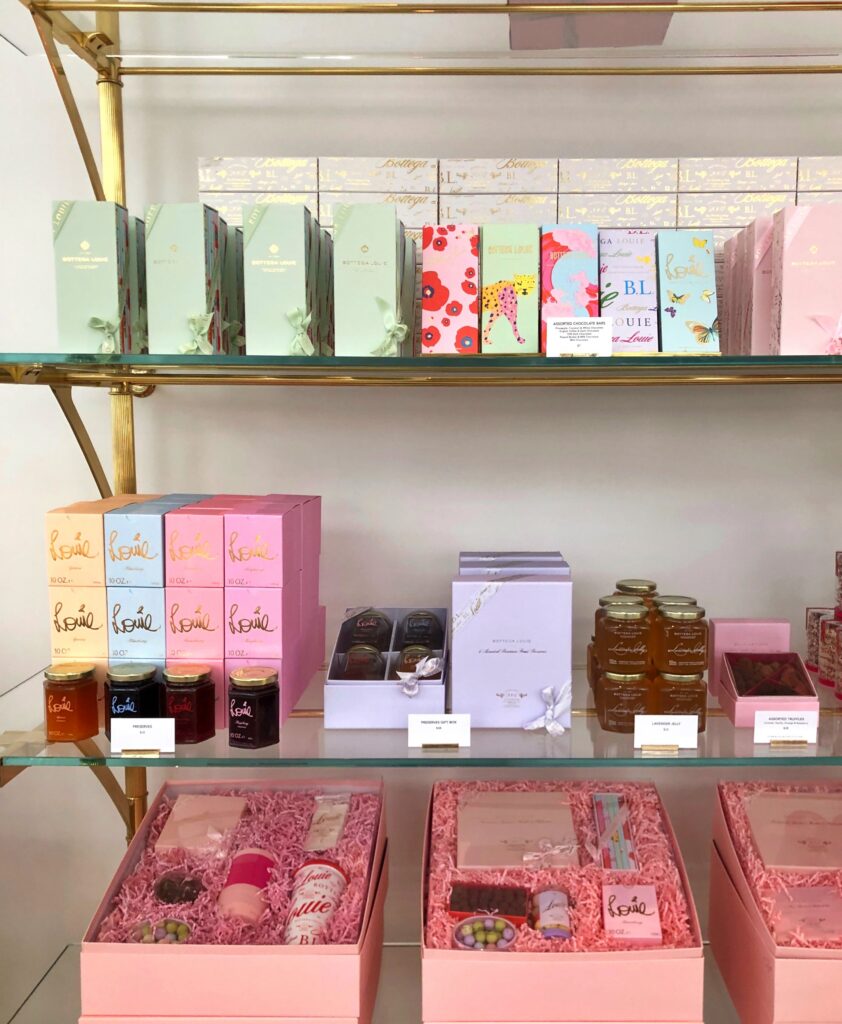 Retail shelves at Bottega Louie in Los Angeles displaying pastel gift boxes, chocolate bars, preserves, honey, truffles, and packaged sweets. Glass shelves, brass supports, pink gift baskets, and colorful product packaging create a polished patisserie-style display.