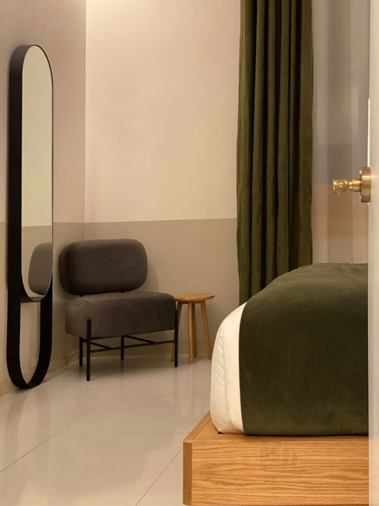 Guest room at the Umbral Curio Hotel in Mexico City with a wood platform bed, dark green bed runner, matching green curtains, and glossy white tile floors. A tall black-framed mirror, gray accent chair, small wood side table, and brass door hardware give the room a simple, modern look.