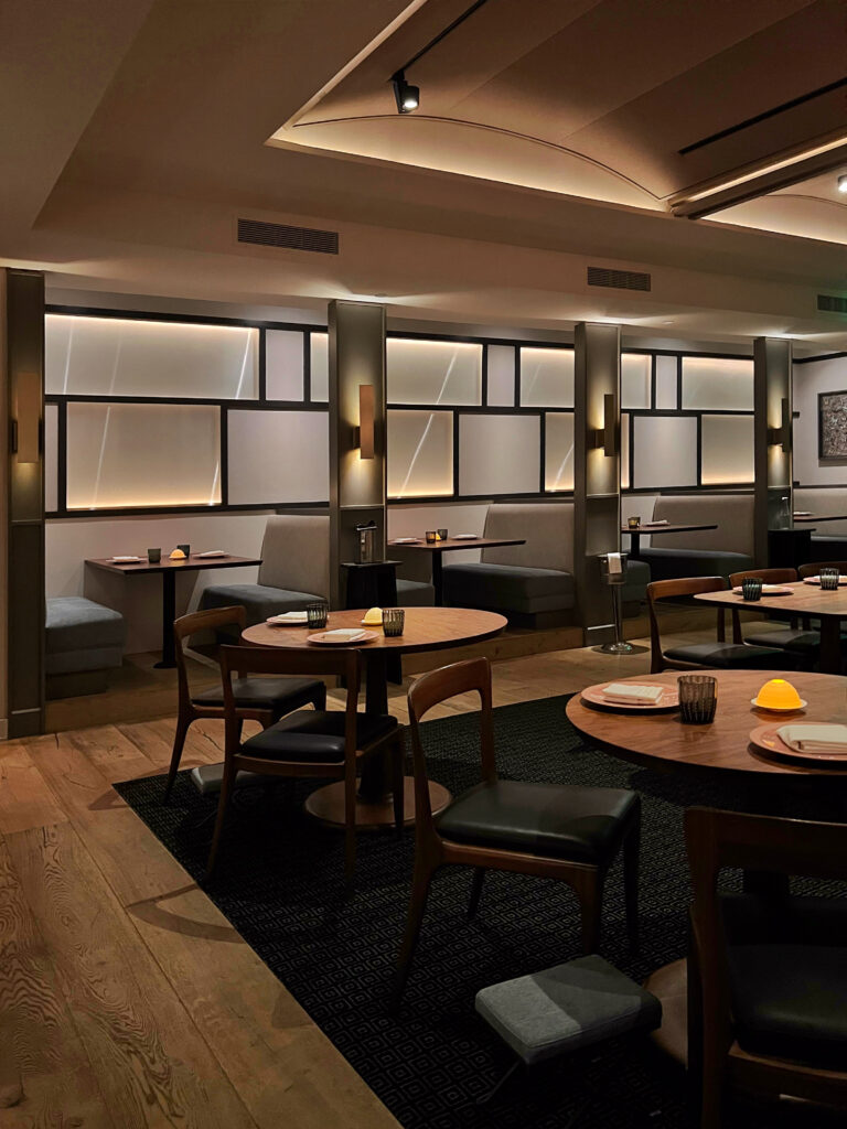 Dining room at Citrin restaurant in Santa Monica with round wood tables, black chairs, gray banquette seating, and small glowing table lamps. Backlit wall panels, wood floors, dark rugs, and warm ceiling lighting give the restaurant a polished, modern atmosphere.