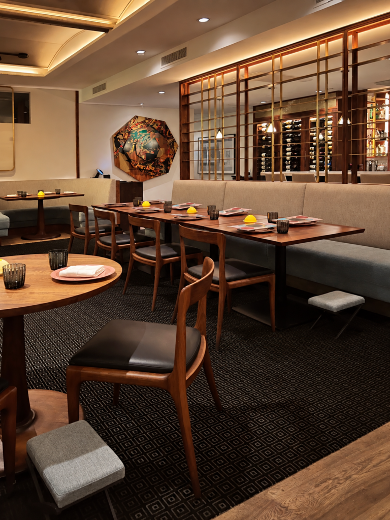 Dining room at Citrin restaurant in Santa Monica with wood tables, gray banquette seating, black patterned carpet, and midcentury-style dining chairs. A glass wine wall, warm ceiling lighting, small table lamps, and colorful wall art add polished detail to the modern restaurant interior.