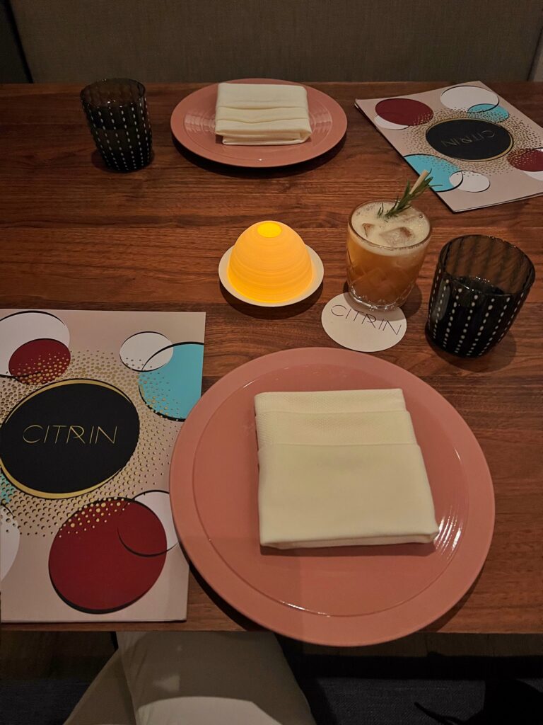Table setting at Citrin restaurant in Santa Monica with peach-colored plates, folded white napkins, patterned menus, dark drinking glasses, and a small glowing table lamp. A cocktail served on a Citrin coaster sits on the wood table between the two place settings.