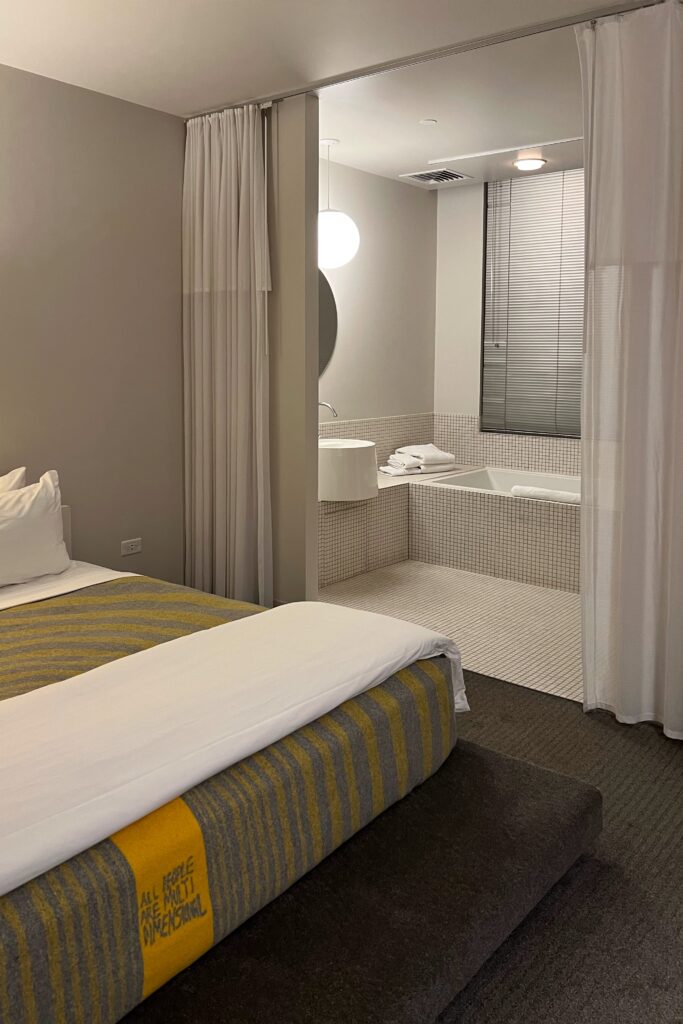 Guestroom at The Delphi Hotel in Los Angeles with a white bed, gray-and-yellow striped blanket, floor-to-ceiling curtain divider, and open bathroom area. The bathroom features an all-white bathroom with mosaic tile, a built-in bathtub, and white towels.