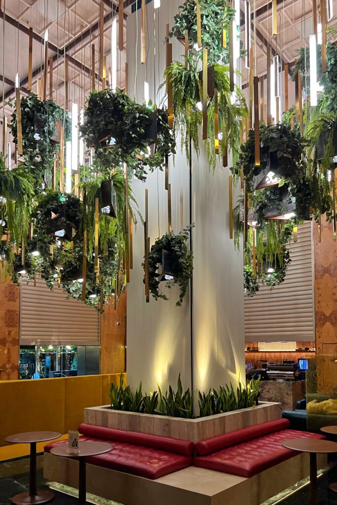 Lobby at The Delphi Hotel in Los Angeles with red tufted banquette seating, small round tables, and a tall central column surrounded by planted greenery. Hanging ferns, trailing plants, mirrors, and vertical pendant lights create a dramatic indoor garden effect above the seating area.