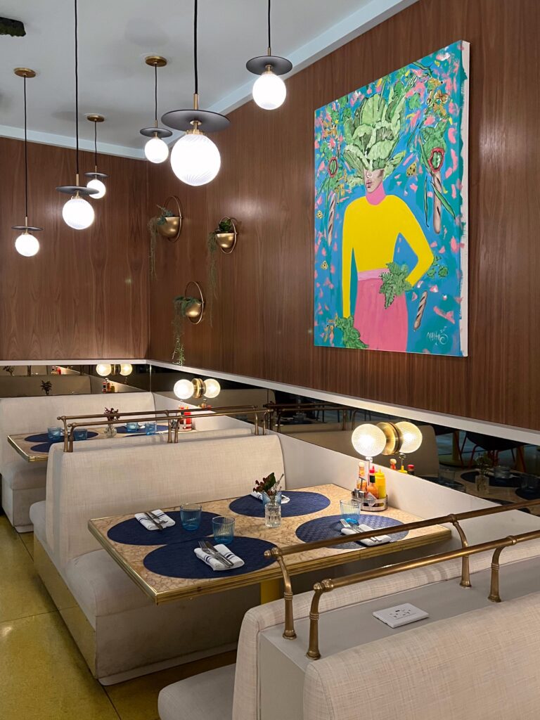 Banquette seating at Denae’s Diner in Los Angeles with cream upholstered booths, gold railings, marble-look tabletops, blue placemats, and glowing globe pendant lights. A colorful pop-art painting hangs on the wood-paneled wall above the dining booths, adding a playful modern detail to the retro diner interior.