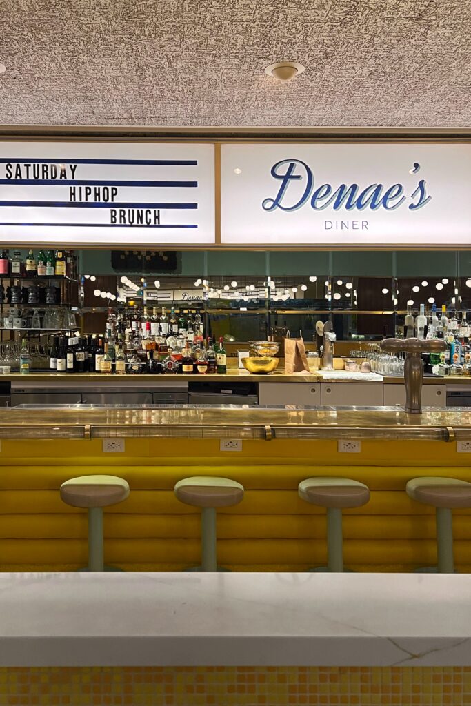 Bar at Denae’s Diner in Los Angeles with a bright yellow counter, round bar stools, liquor bottles, mirrored back wall, and an illuminated Denae’s Diner sign. A marquee-style sign reading “Saturday Hip Hop Brunch” adds a retro diner detail above the bar.