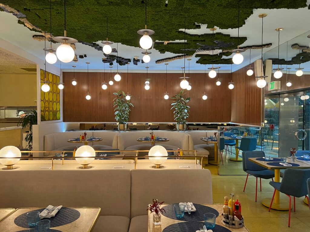 Wide view of Denae’s Diner in Los Angeles with cream banquettes, gold-trimmed tables, blue dining chairs, yellow floors, and rows of globe pendant lights. The green textured ceiling installation, wood-paneled walls, potted plants, and retro yellow artwork give the dining room a colorful, modern diner look.