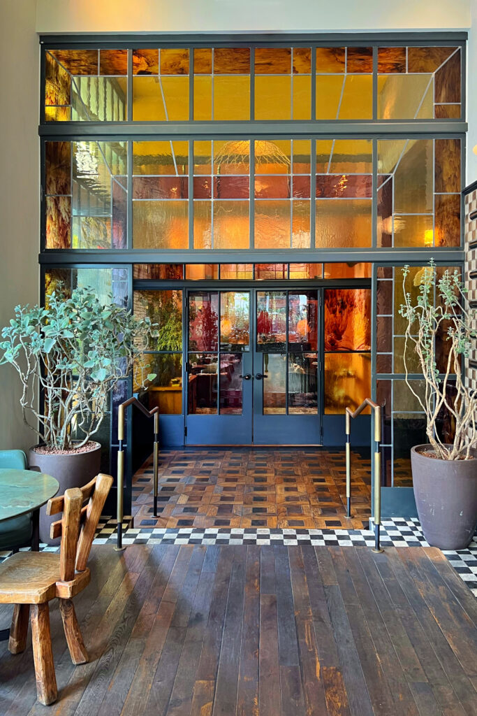 Interior at the Los Angeles Proper Hotel with tall black-framed stained glass doors, warm amber glass panels, potted plants, and checkerboard tile flooring. Wood floors, brass handrails, rustic chairs, and textured glass create a layered entryway leading into the restaurant space.