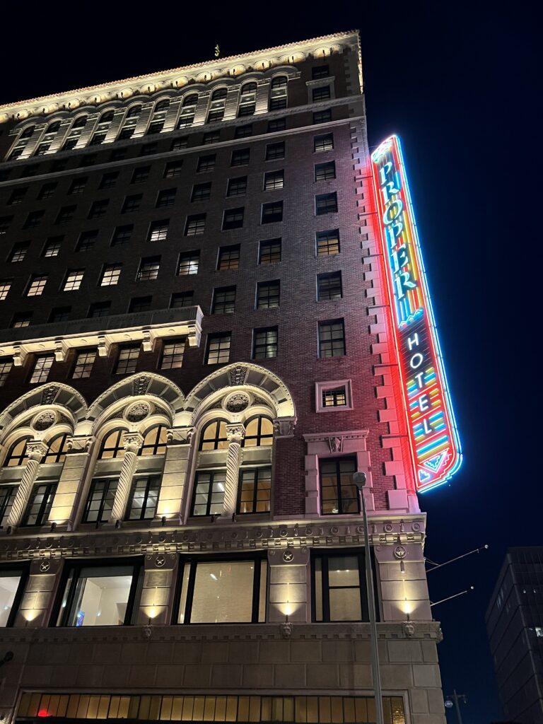 Exterior of the Los Angeles Proper Hotel at night, showing the historic brick facade, illuminated arched windows, ornate architectural details, and vertical neon Proper Hotel sign. The red, blue, yellow, and white neon sign glows against the dark sky along the side of the building.