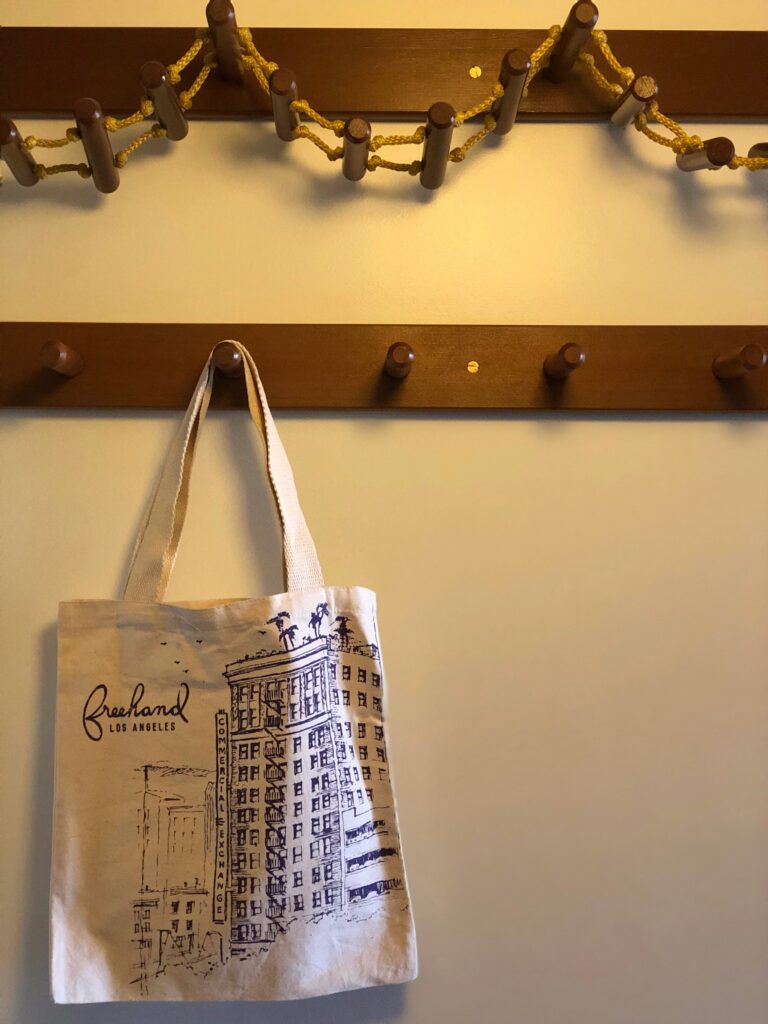 Canvas Freehand Los Angeles tote bag hanging from a wooden wall hook inside a hotel guestroom. Above the bag, a decorative rope-and-wood wall rack adds a warm, playful detail to the room’s simple wall storage.