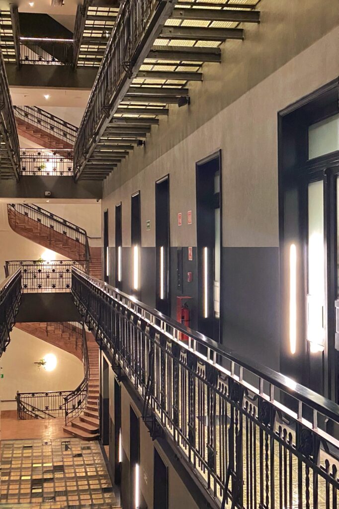 Interior atrium at Hotel Umbral Curio in Mexico City with black metal balcony railings, illuminated walkways, and a multi-level staircase. The view shows tall guestroom doors, gray walls, warm wall lights, and patterned glass floor panels inside the historic hotel building.