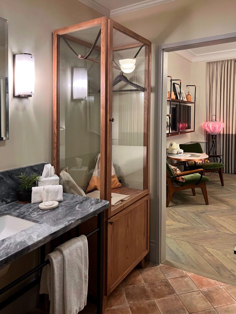 Guestroom vanity and wardrobe at The Hoxton Hotel Downtown Los Angeles with a gray marble vanity, wall sconces, terracotta floor tile, and a glass-front wood wardrobe beside the sink. Through the doorway, the guest room shows herringbone wood floors, green lounge chairs, a small round table, shelving, draped curtains, and a pink sculptural floor lamp.