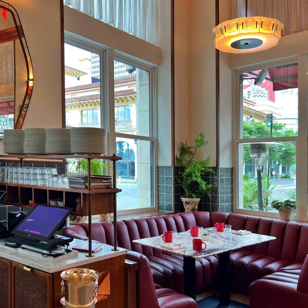 Restaurant seating area at The Hoxton Downtown Los Angeles with a curved red leather banquette, marble table, red mugs, stacked plates, and tall windows facing the street. The space features glossy green tile, warm wood trim, indoor plants, a sculptural ceiling light, and a polished boutique hotel dining room look.