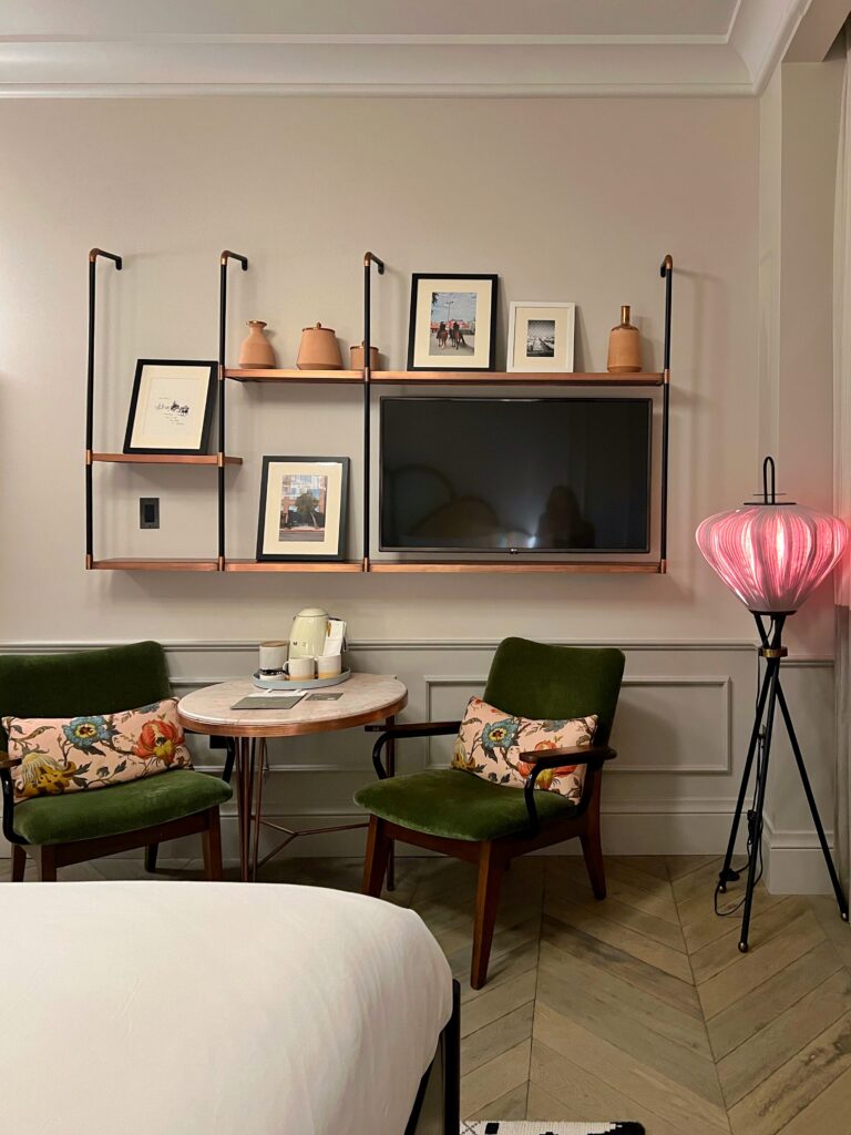 Guest room at The Hoxton Downtown Los Angeles with two green velvet chairs, floral pillows, a small round table, wall-mounted shelving, framed art, and a television. A pink sculptural floor lamp, pale paneled walls, herringbone wood floors, and the edge of a white bed add color and texture to the boutique hotel room.