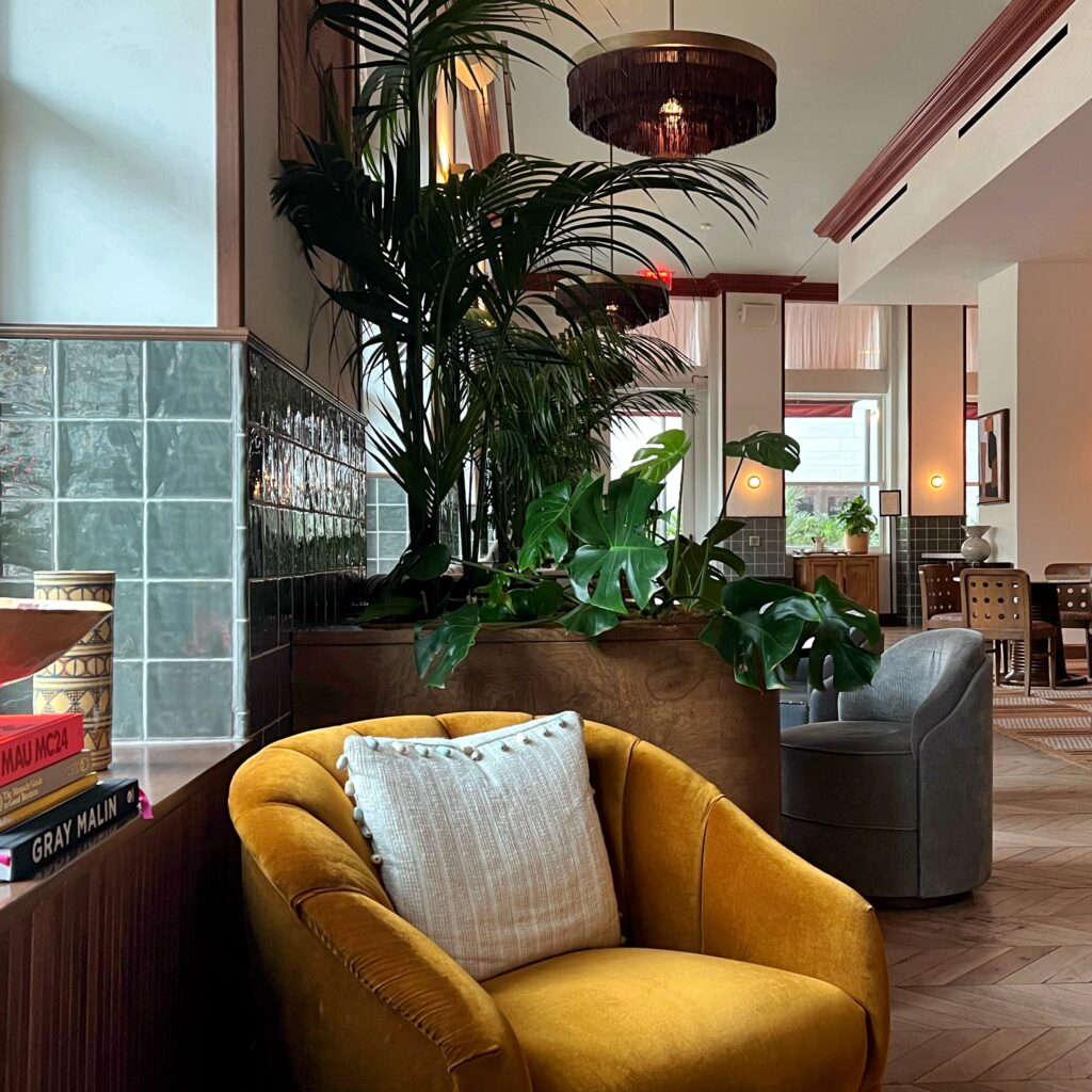 Lobby seating area at The Hoxton Downtown Los Angeles with a mustard velvet chair, white textured pillow, large indoor plants, glossy green tile, and wood accents. The space includes herringbone wood floors, upholstered lounge chairs, books, warm wall sconces, and a layered boutique hotel interior in soft natural light.