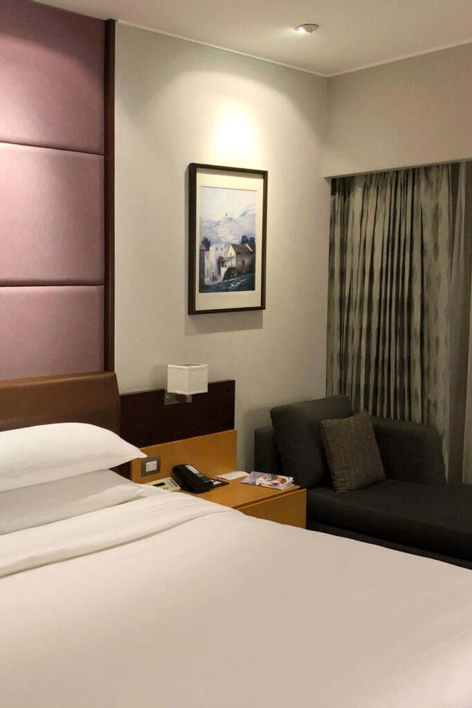 Guest room at Hyatt Regency Mexico City with a white bed, tall upholstered headboard, bedside table, framed artwork, and gray sitting chair by the window. Patterned curtains, warm bedside lighting, and neutral walls give the hotel room a simple, polished look.