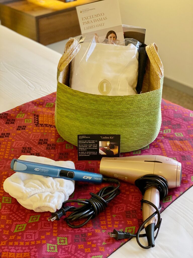 Women’s kit in a Ladies Suite at InterContinental Polanco with a green woven basket, packaged amenities, a hair dryer, flat iron, and white cloth accessory arranged on the bed. The items sit on a bright pink patterned textile runner, adding color and detail to the hotel room amenity setup.