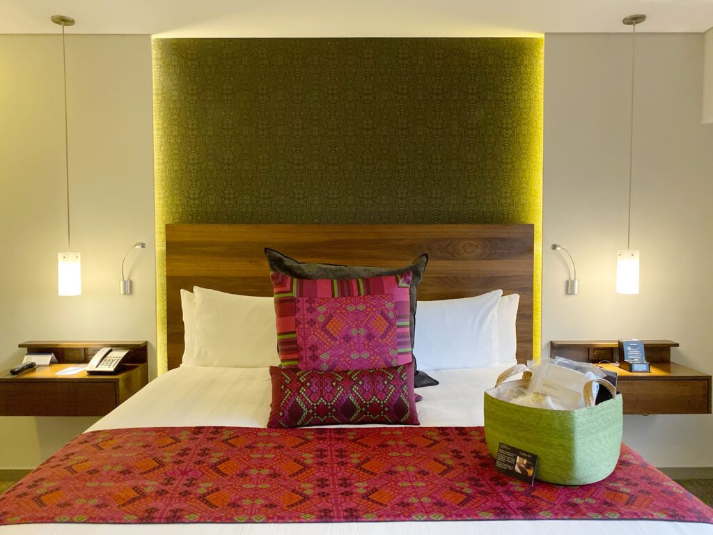 Where to stay in Polanco Mexico City. This is the Guest room at InterContinental hotel with a wood headboard, green patterned wall panel, white bedding, and colorful pink-and-red textiles layered across the bed. Floating bedside tables, pendant lights, reading sconces, and a green welcome basket add detail to the modern hotel room.