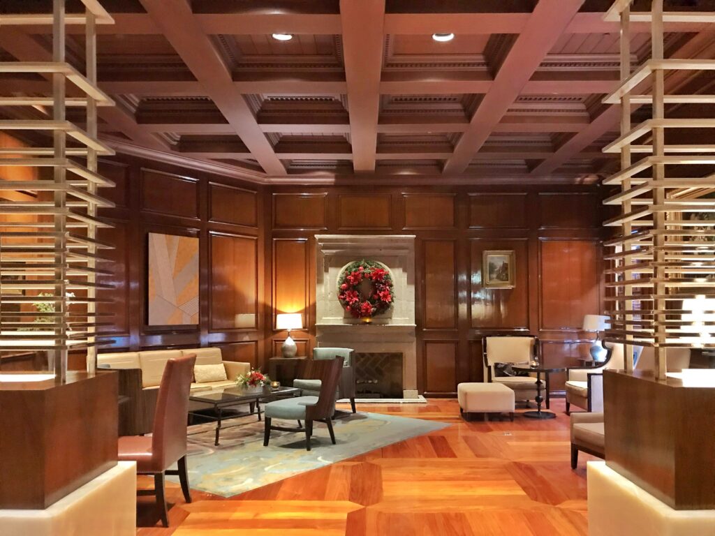 Lobby lounge at JW Marriott Mexico City with wood-paneled walls, a coffered ceiling, parquet wood floors, and a stone fireplace decorated with a floral wreath. Upholstered chairs, sofas, table lamps, framed artwork, and warm lighting create a classic hotel seating area.