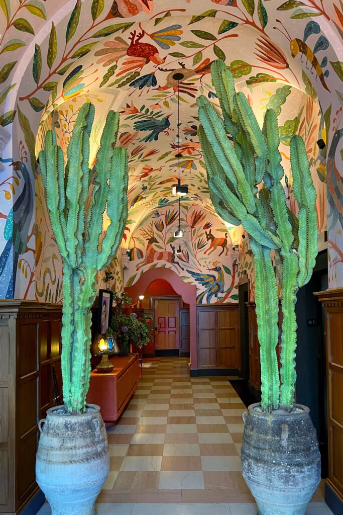 Hallway at the Los Angeles Proper Hotel, designed by Kelly Wearster, with two tall cactus planters, wood wall paneling, checkerboard tile flooring, and an arched ceiling covered in a colorful botanical mural. Painted birds, flowers, leaves, and playful animal motifs wrap across the walls and ceiling, creating a vibrant entryway inside the hotel.