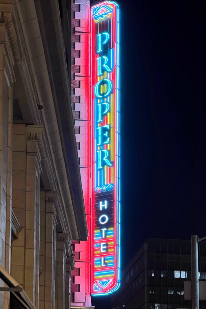 Exterior view of the Los Angeles Proper Hotel neon sign glowing in bright red, blue, yellow, and white against the night sky. The vertical sign is mounted on the side of the historic hotel building, with downtown Los Angeles buildings visible in the background.