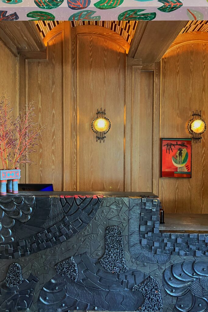 Reception area at the Los Angeles Proper Hotel with a sculptural black front desk, warm wood-paneled walls, round wall sconces, and colorful artwork. A painted botanical mural continues across the ceiling edge above the desk, adding color and pattern to the hotel entry.