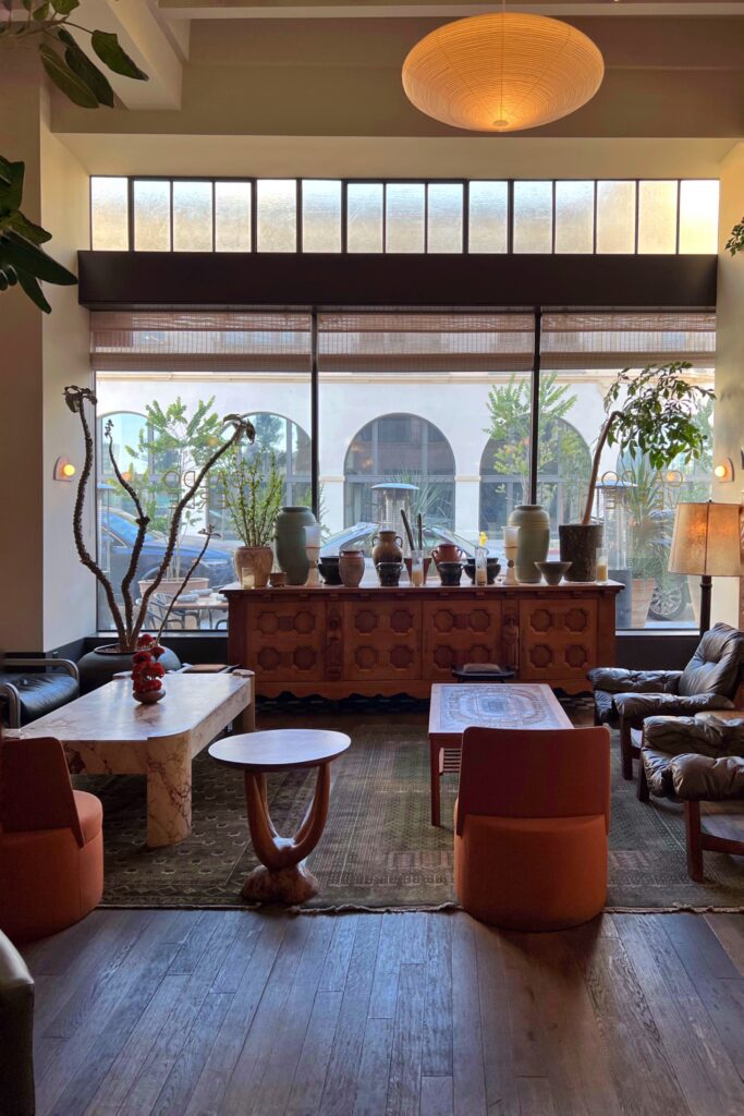 Lobby lounge at the Los Angeles Proper Hotel with tall windows, potted plants, vintage-style leather chairs, orange lounge seating, patterned rugs, and a carved wood console. A large round pendant light, marble coffee table, ceramic vessels, and warm natural light give the seating area a layered, collected look.