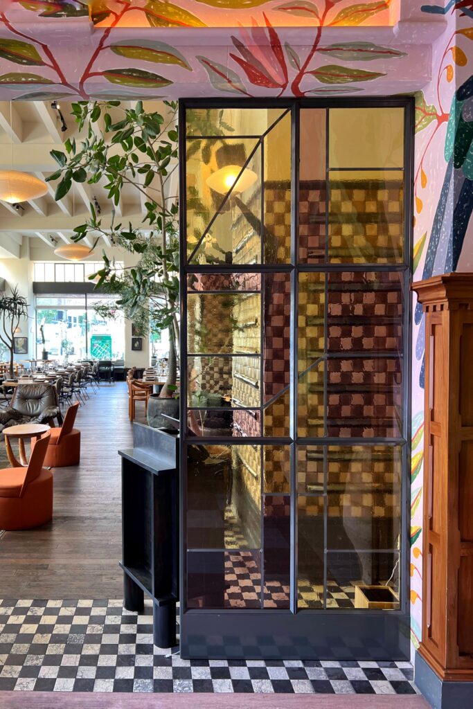Interior at the Los Angeles Proper Hotel, designed by Kelly Wearstler, with a tall black-framed stained glass stairwell, checkerboard tile flooring, wood paneling, and colorful painted botanical murals across the surrounding walls and ceiling. The view opens into a bright restaurant space with trees, pendant lights, lounge chairs, and dining tables in the background.
