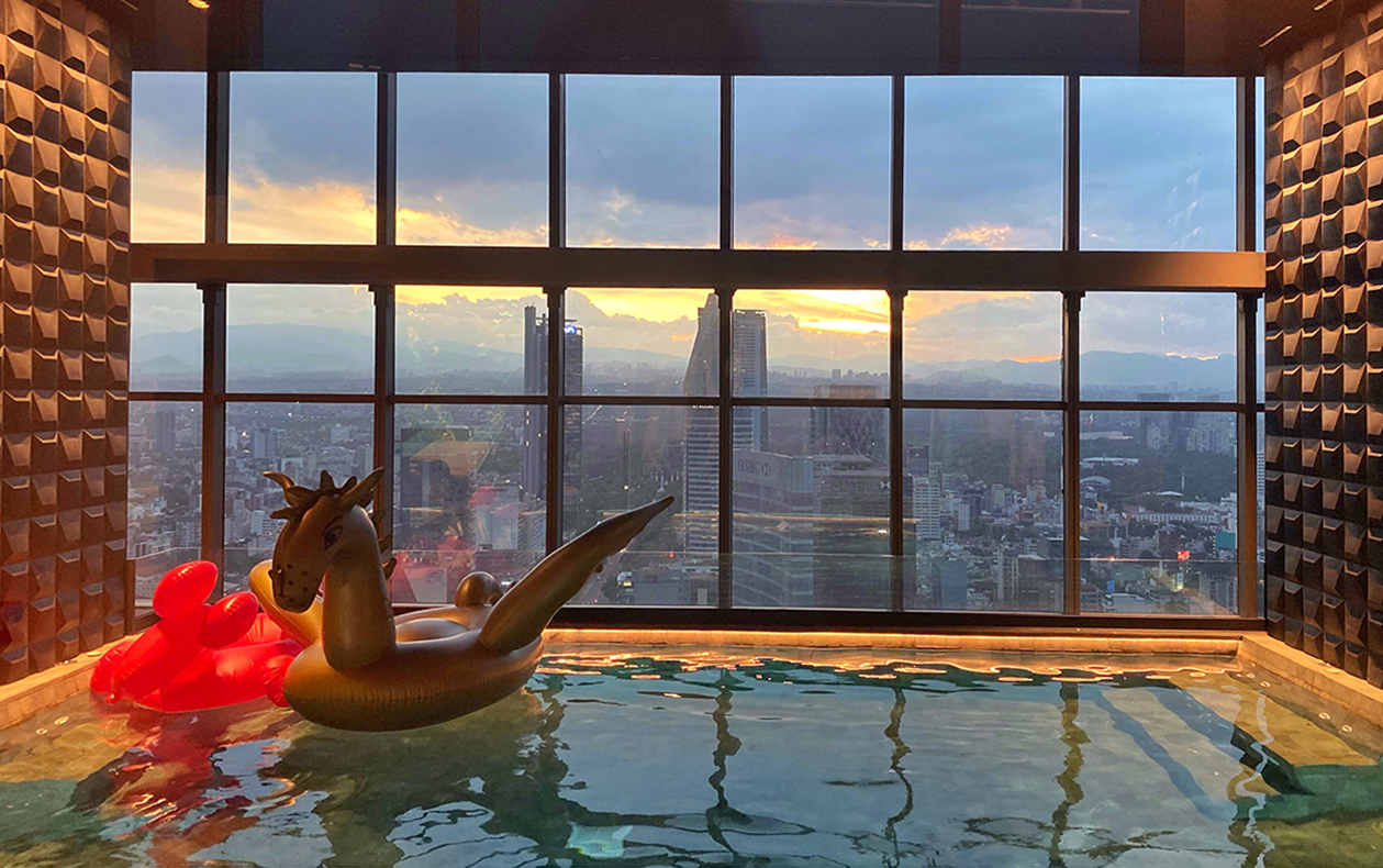 Indoor rooftop pool with floor-to-ceiling windows overlooking the Mexico City skyline at sunset. A gold unicorn pool float and red inflatable float sit in the water, with geometric textured walls and warm lighting framing the city view.