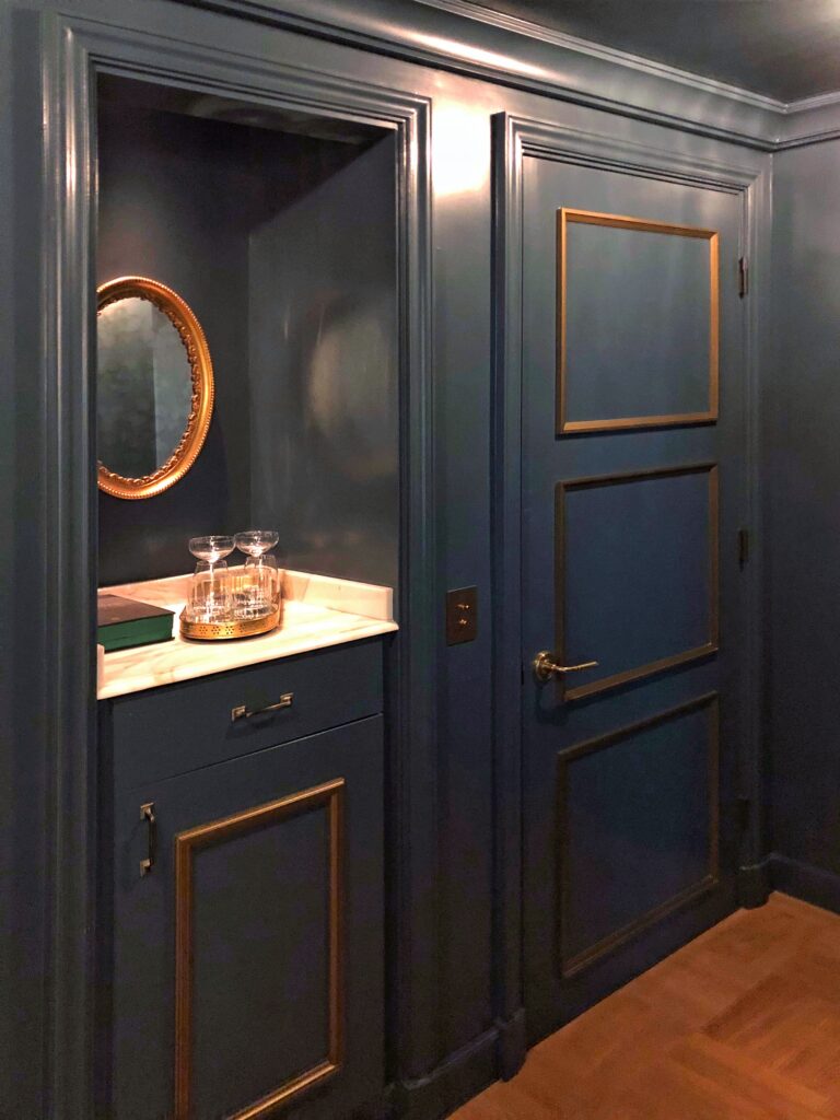 Color-drenched guest room at Per La Hotel in Los Angeles with glossy deep teal walls, matching paneled door, brass trim, and warm wood flooring. A built-in marble-topped niche holds glassware, a tray, and a round gold-framed mirror illuminated by soft ambient light.