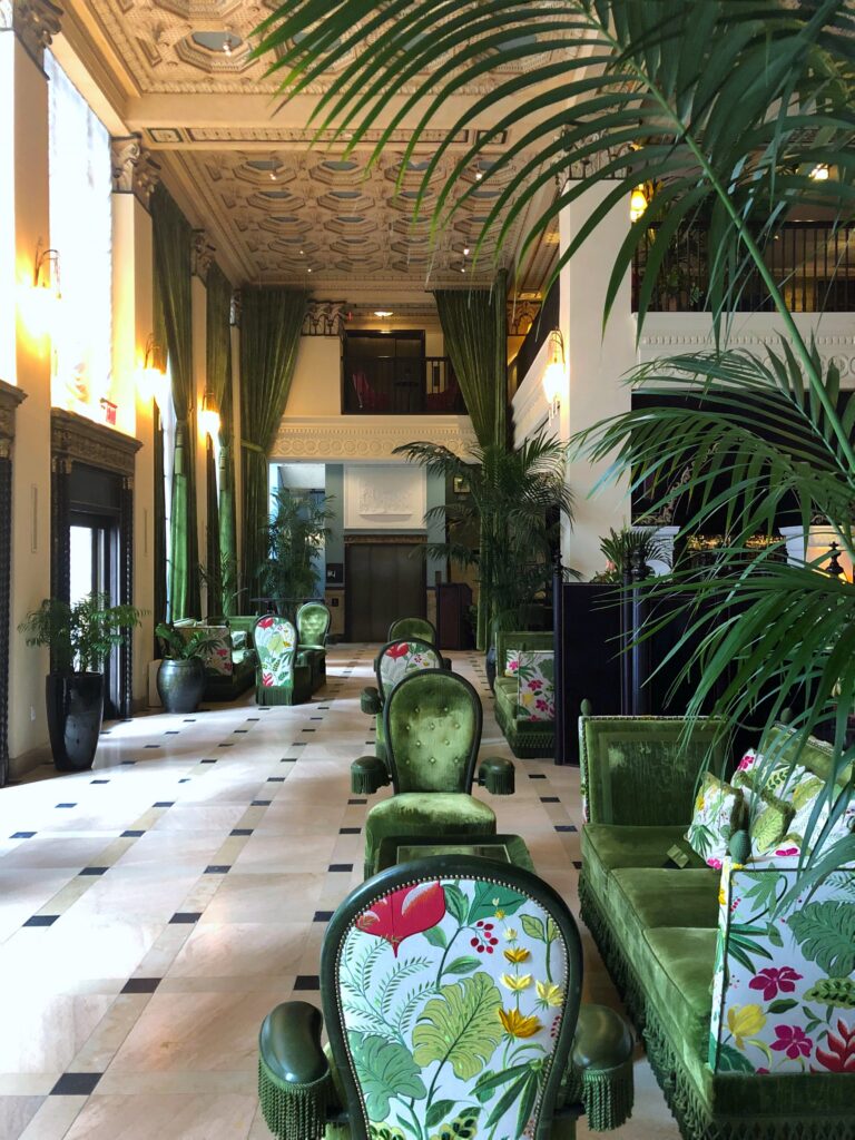 Lobby at Per La Hotel in Los Angeles with green velvet chairs, floral upholstery, palm plants, tall green curtains, and patterned marble flooring. The space features an ornate coffered ceiling, warm wall sconces, high columns, and layered tropical-inspired seating.