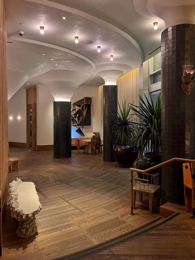 Lobby at Proper Hotel Santa Monica with dark tiled columns, sculptural plaster ceiling forms, warm uplighting, and patterned wood flooring. Potted plants, sculptural seating, wall art, and layered wood details add texture to the hotel’s design-forward interior.