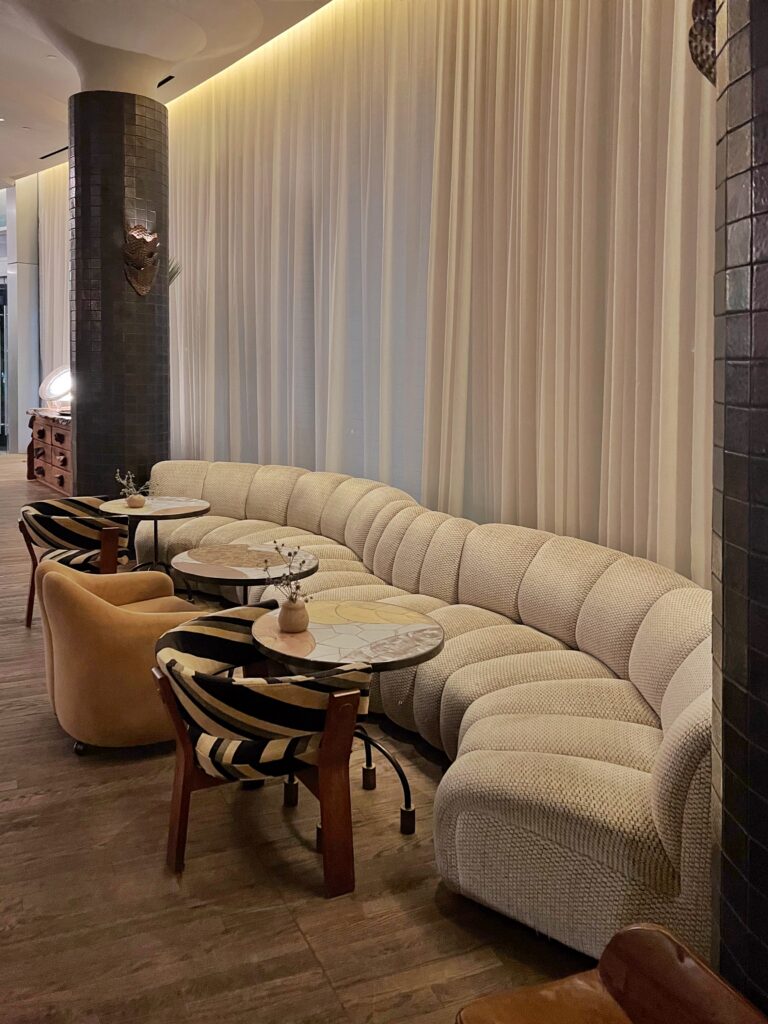 Lobby lounge at Proper Hotel Santa Monica with a long curved cream banquette, layered round tables, striped accent chairs, and a mustard upholstered lounge chair. Floor-to-ceiling drapery, dark tiled columns, warm lighting, and wood flooring create a soft, design-forward hotel seating area.