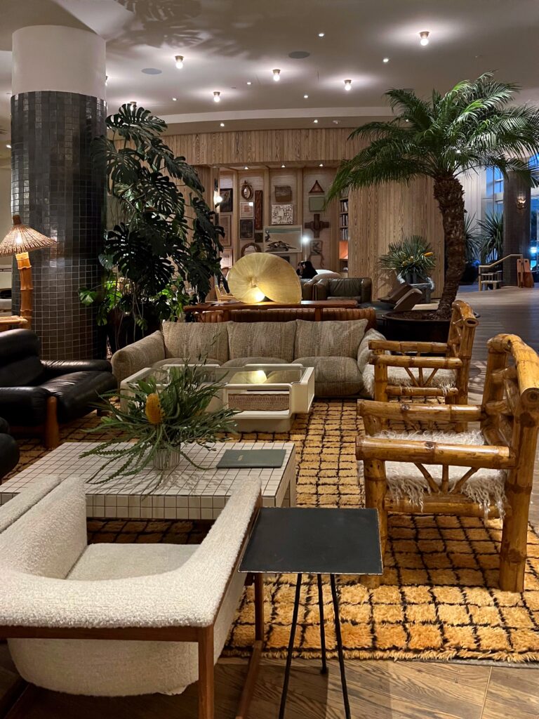 Lobby lounge at Proper Hotel Santa Monica with layered seating, including cream upholstered chairs, leather sofas, rustic wood armchairs, and low coffee tables. Palm trees, large potted plants, dark tiled columns, wood-paneled walls, and a patterned yellow rug create a warm, collected hotel lobby interior.